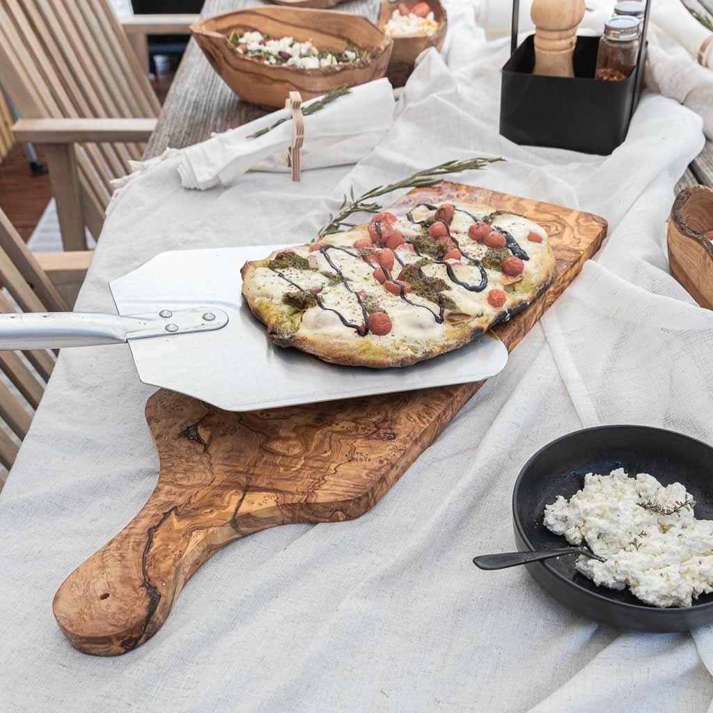 Pizzaschieber Classic Personal Edition mit ergonomischem Holzgriff und Edelstahlfläche neben frischer Pizza auf hölzernem Tisch.