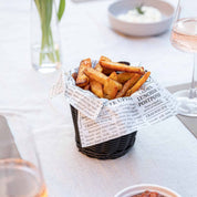 Pommes in einem stilvollen Pommeskörbchen mit Zeitungspapier auf einem gedeckten Tisch serviert.