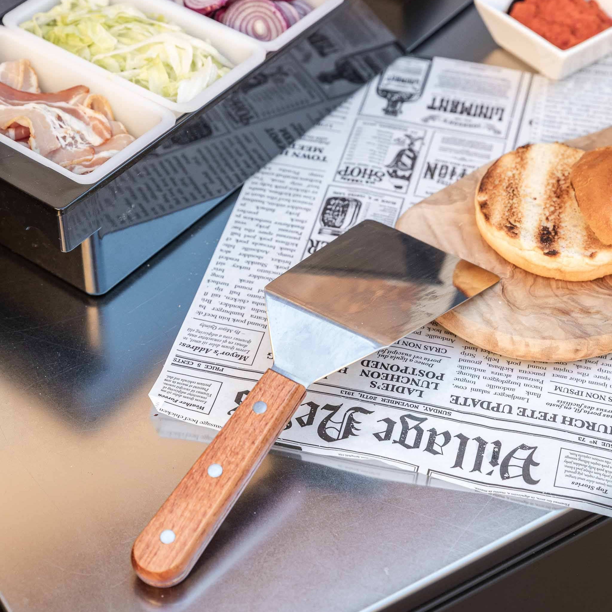 Pfannenwender mit Edelstahlfläche und Holzgriff auf Küchentheke neben Zutaten.